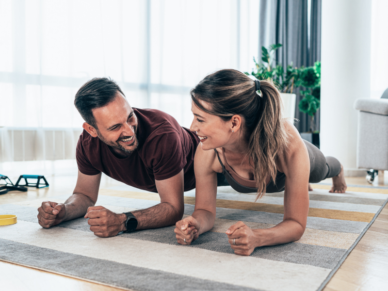 A man and woman smile at each other while doing plank exercises on a rug in a bright living room.