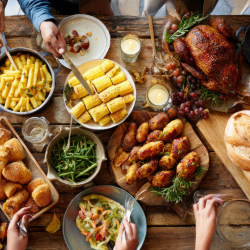 A wooden table set with roasted chicken, corn on the cob, salad, bread, green beans, pasta, roasted potatoes, and people serving themselves food.