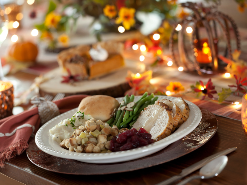 A Thanksgiving dinner plate with turkey, stuffing, green beans, cranberry sauce, mashed potatoes, and a roll on a festive table with autumn decorations and lights.