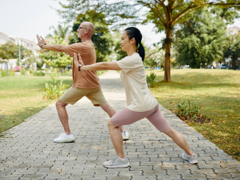 Two people practicing tai chi outdoors in a park, standing in a lunge position with arms extended forward on a paved path surrounded by greenery.