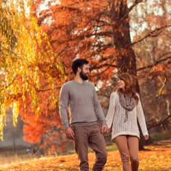 A couple holding hands and smiling while walking through a park with vibrant autumn trees and fallen leaves.