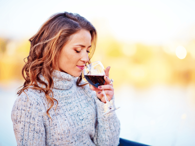 When You Reach For Alcohol, Are You Actually Looking For A Boost Of GABA? - A woman with wavy hair in a gray sweater gently holds and smells a glass of red wine, enjoying the aroma outdoors as she savors a relaxing gaba moment.