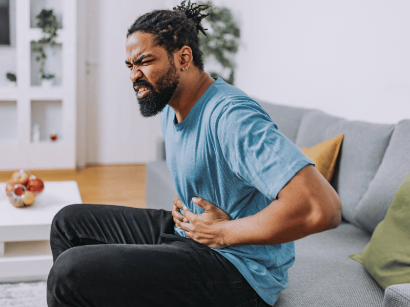 A man sitting on a couch clutching his stomach and grimacing in pain, possibly experiencing discomfort from an h pylori infection.