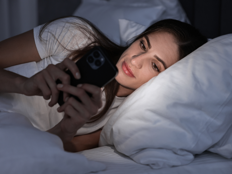 A woman lies in bed at night, propped on a pillow, using her smartphone and smiling softly. The glow of the phone lights up her face in the dimly lit room.