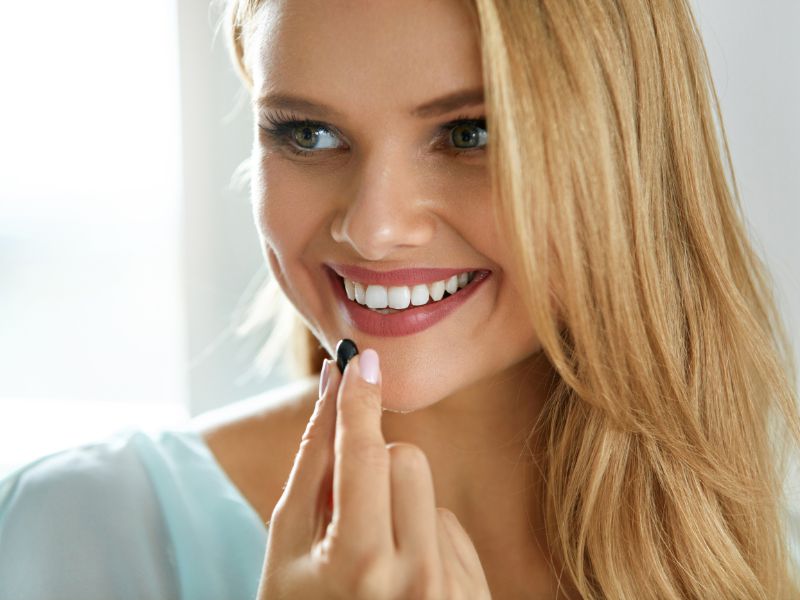 A smiling woman with long blonde hair holds a small dark pill or supplement near her mouth, ready to take it.