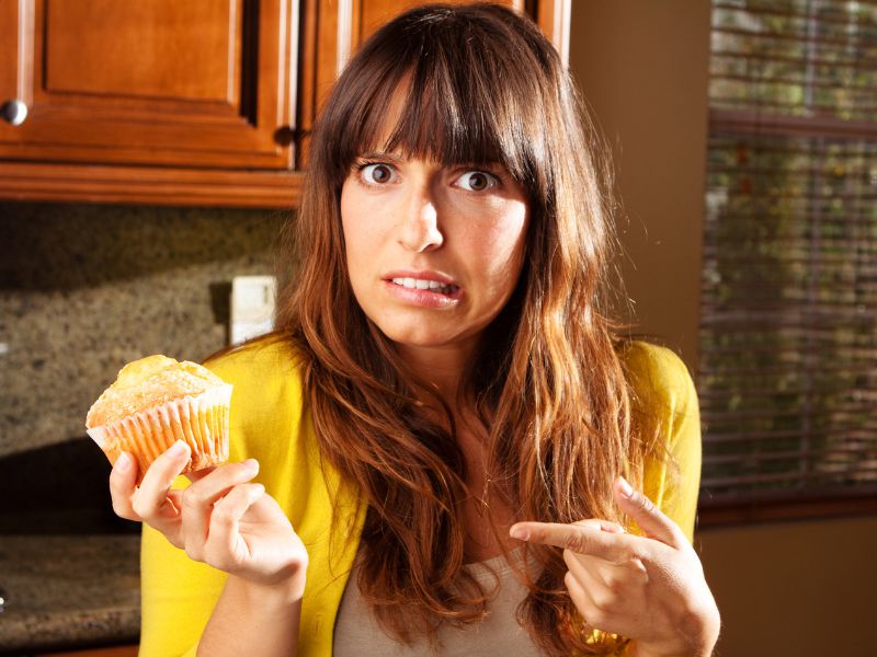 A woman with long brown hair and a yellow sweater looks confused and points at a muffin she is holding in a kitchen.