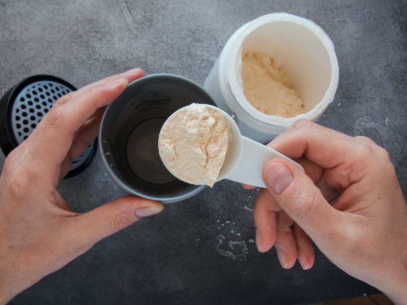 A person prepares a protein shake, holding a scoop of protein powder above a shaker bottle with an open container of powder nearby on a grey surface.