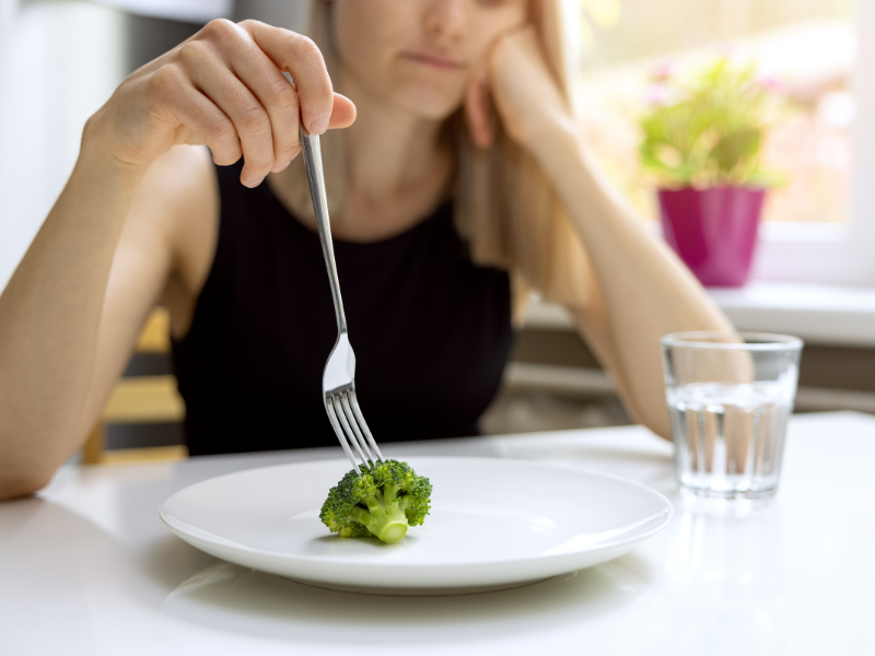 A woman sits at a table, looking bored, holding a fork with a lone piece of broccoli on an otherwise empty plate. A glass of water rests beside her.