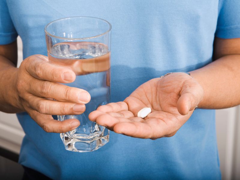 A person in a blue shirt holds a glass of water in one hand and a white pill in the other hand.