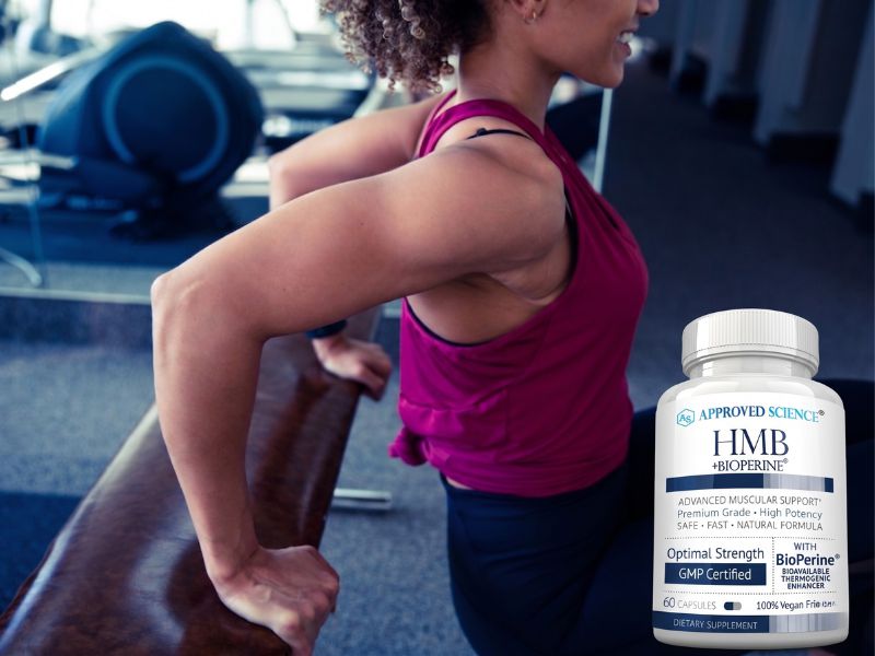 A woman in workout clothes does tricep dips in a gym. In the foreground, there is a bottle of Approved Science HMB + BioPerine supplement.