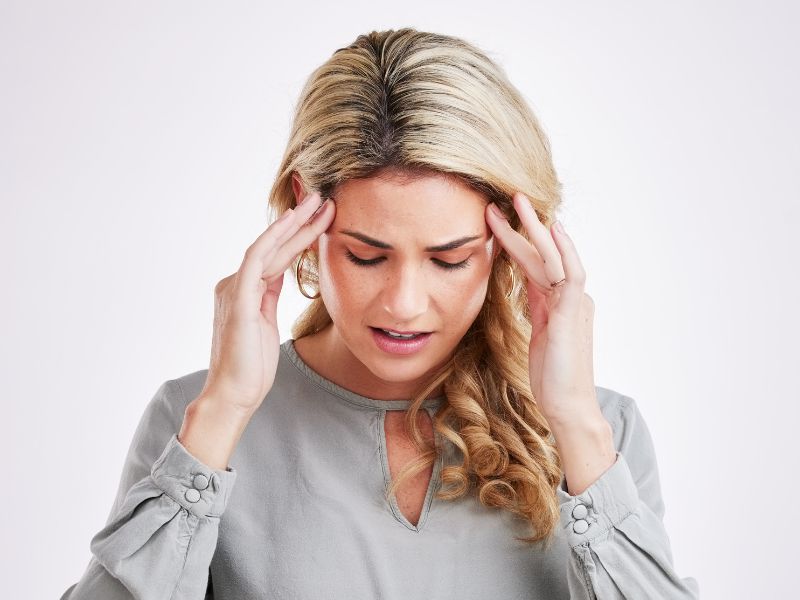 A woman with blonde hair touches her temples with both hands and looks down, appearing to have a headache or be experiencing brain fog.