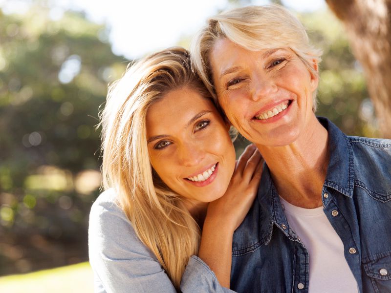 Two women smiling outdoors, close together, with one woman resting her head on the others shoulder. Both have light hair and are wearing casual denim shirts. Trees blurred in the background.