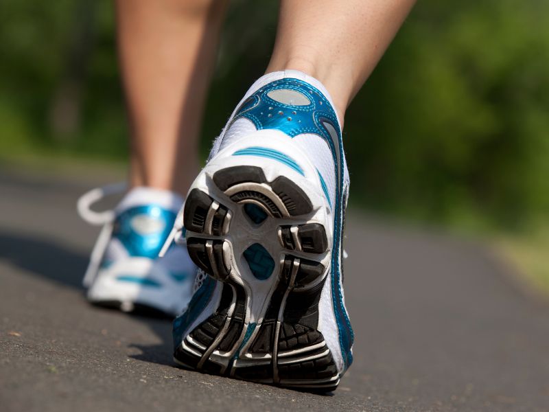 Close-up of a person walking on a paved path, showing the sole of a white and blue athletic shoe with a blurred green background.