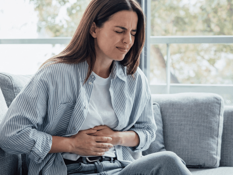 A woman sitting on a couch holds her stomach and appears to be in pain or discomfort, suggesting she might have a stomachache.